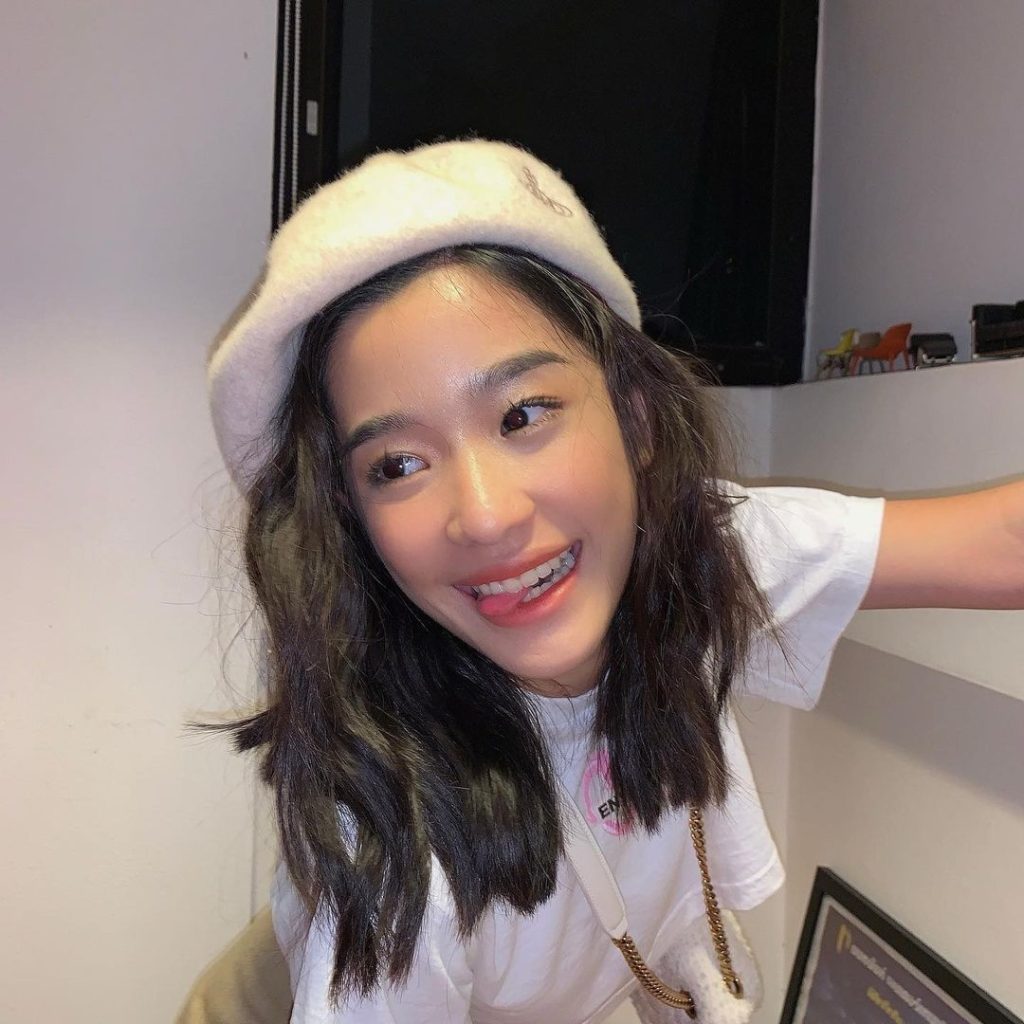 Tu Tontawan wearing a beanie and white shirt, with a bit of her tongue out her mouth and smiling.