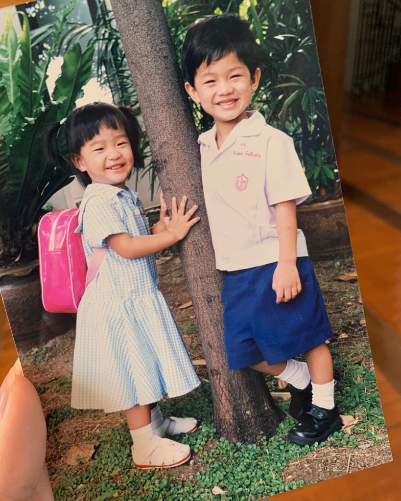 A young Tu Tontawan and her brother posing next to a tree and smiling for the camera. Tu has both her hands on the tree.