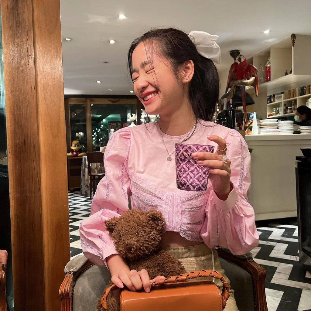 A celebrity wearing a pink shirt with sleeves, turning her head to the side and laughing at a cafe.