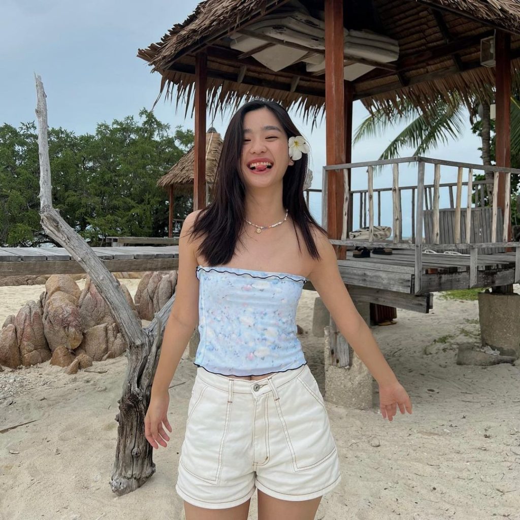 A celebrity with a flower in her hair, sleeveless crop top and shorts on the beach.