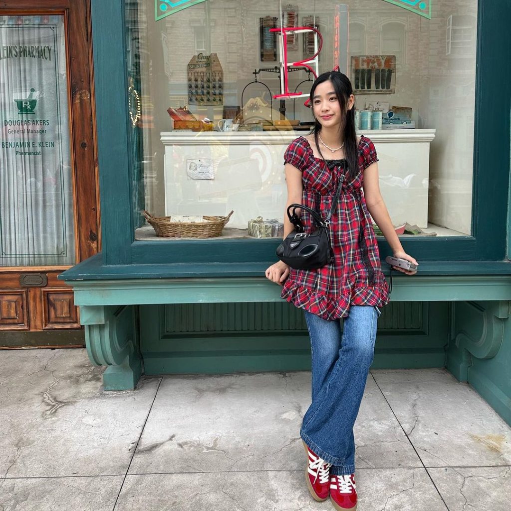 Tu Tontawan wearing a red plaid dress and jeans, sitting on a window ledge outside a shop.