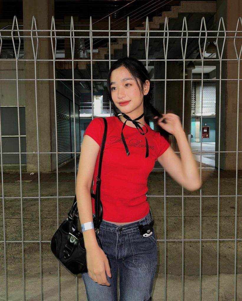 Tu Tontawan Thai Actress wearing a red shirt with her hand touching her hand, standing in front of a metal grill door.