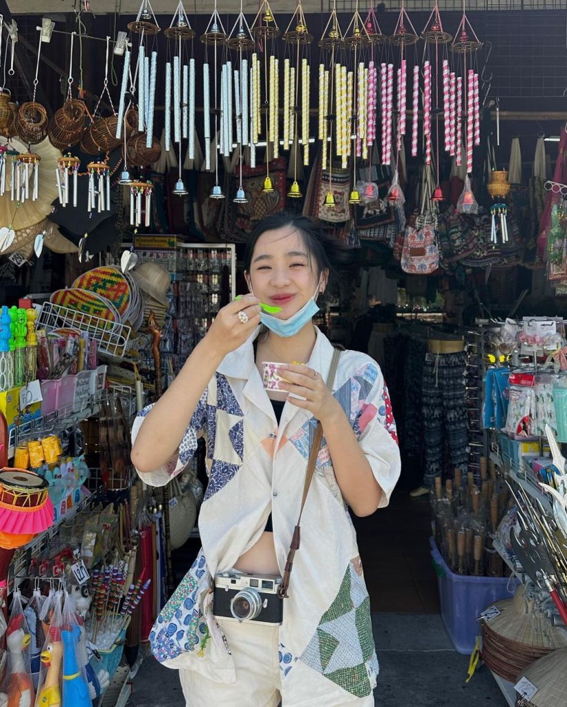 Tu Tontawan wearing a patchwork top, eating ice cream and smiling in front of a traditional shop.