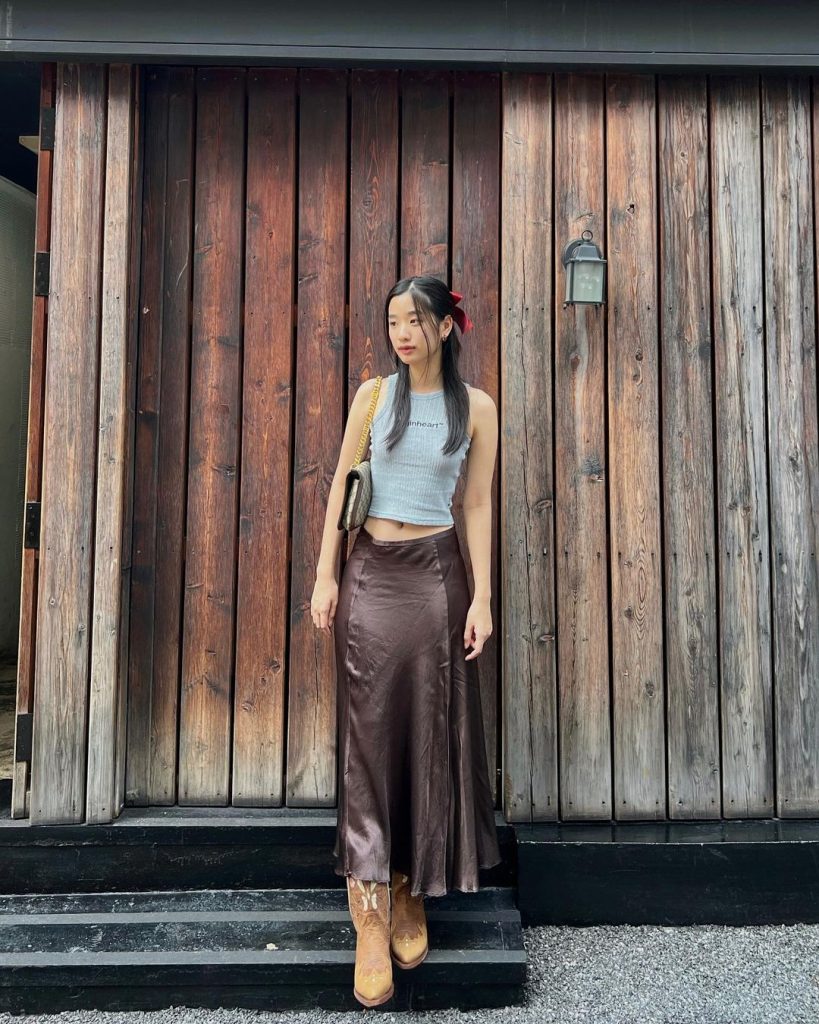 Tu Tontawan wearing a grey crop top and low waisted brown leather skirt, posing in front of a wooden wall.